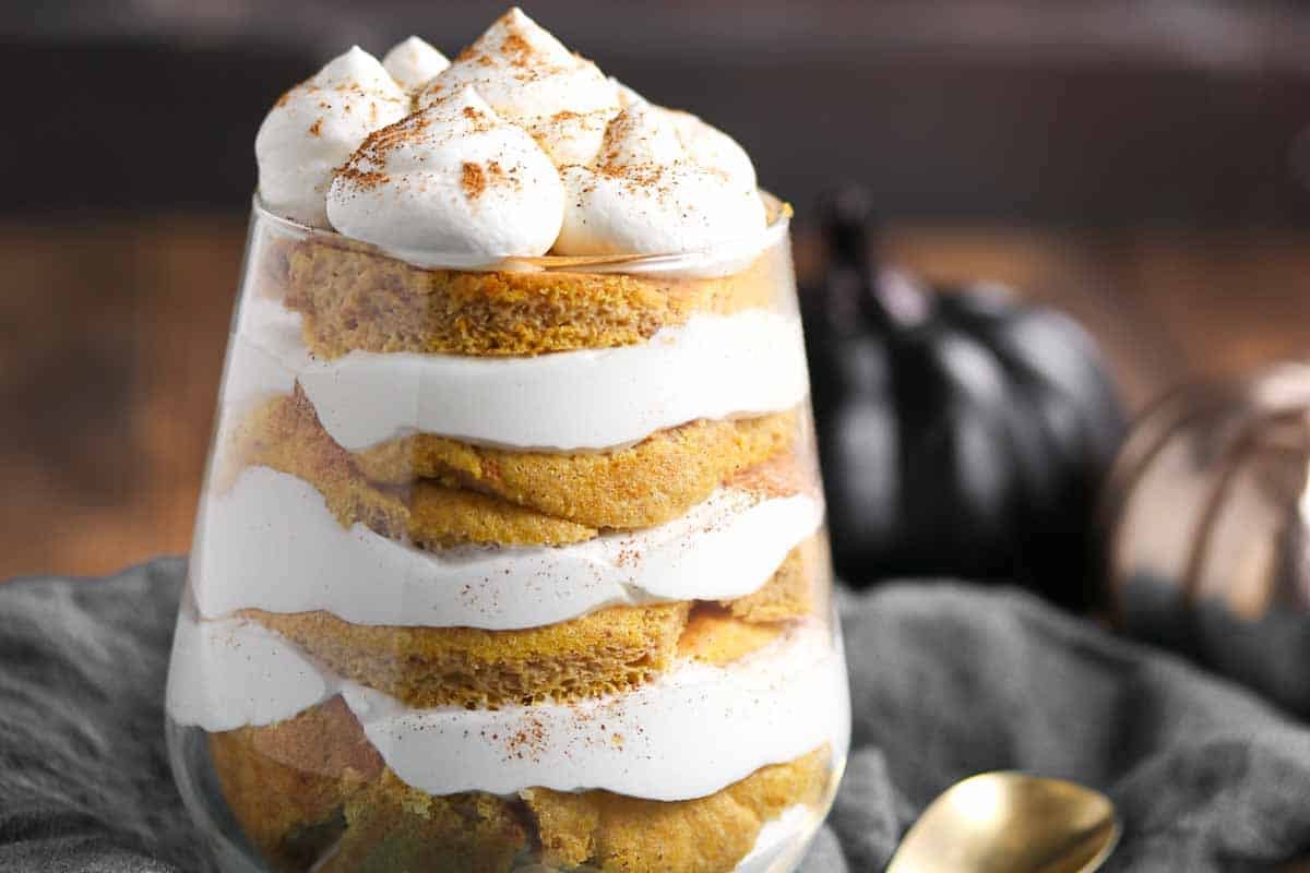A glass layered with pumpkin cake and whipped cream, topped with piped cream and a dusting of cinnamon.
