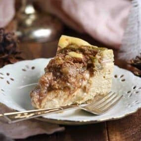 A slice of apple streusel cheesecake on a decorative white plate with a fork.