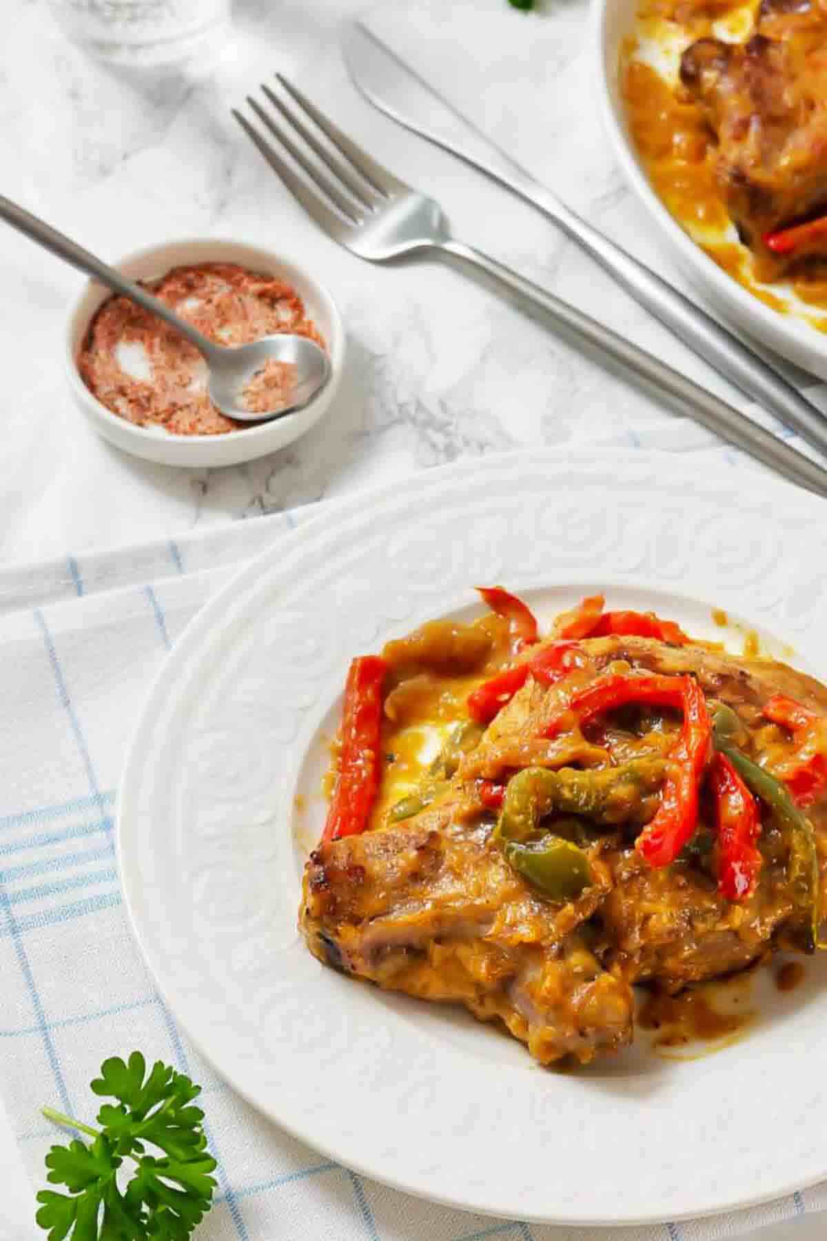Chicken cacciatore with red and green peppers on a white plate, with a small dish of seasoning and cutlery nearby.
